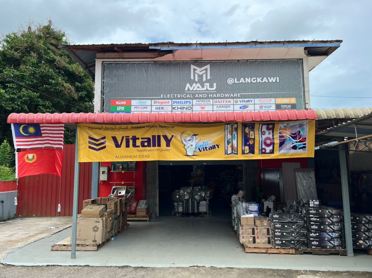 Hardware shop with a dark MAJU @LANGKAWI signboard and a row of brand logos, a yellow Vitally banner, and boxed fans and Deka water heaters stacked on pallets outside