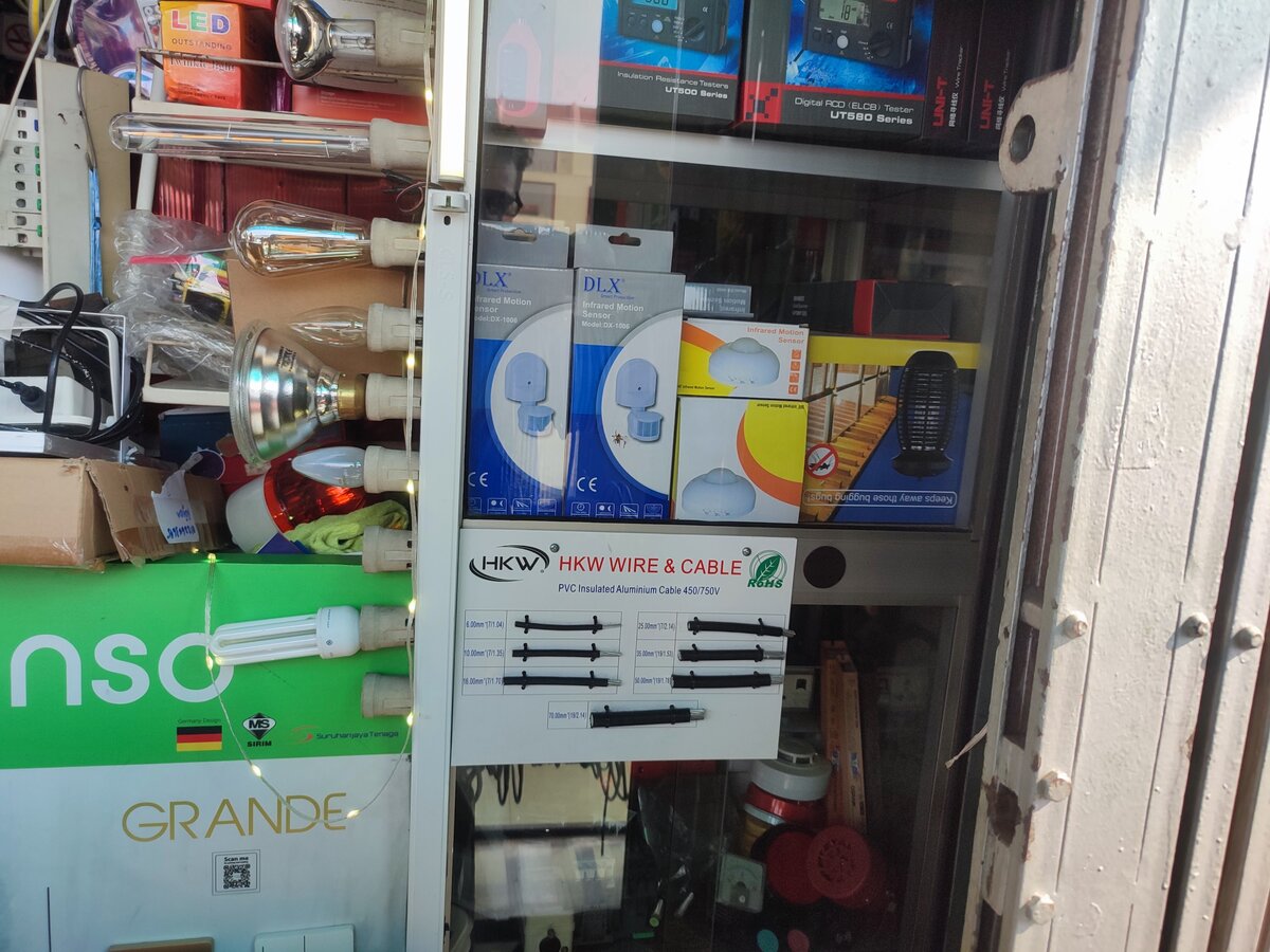 Glass display cabinet with a white board of black aluminium cable samples labelled HKW Wire and Cable, boxes of UNI-T testers and DLX motion sensors, and light bulbs in lampholders on the left