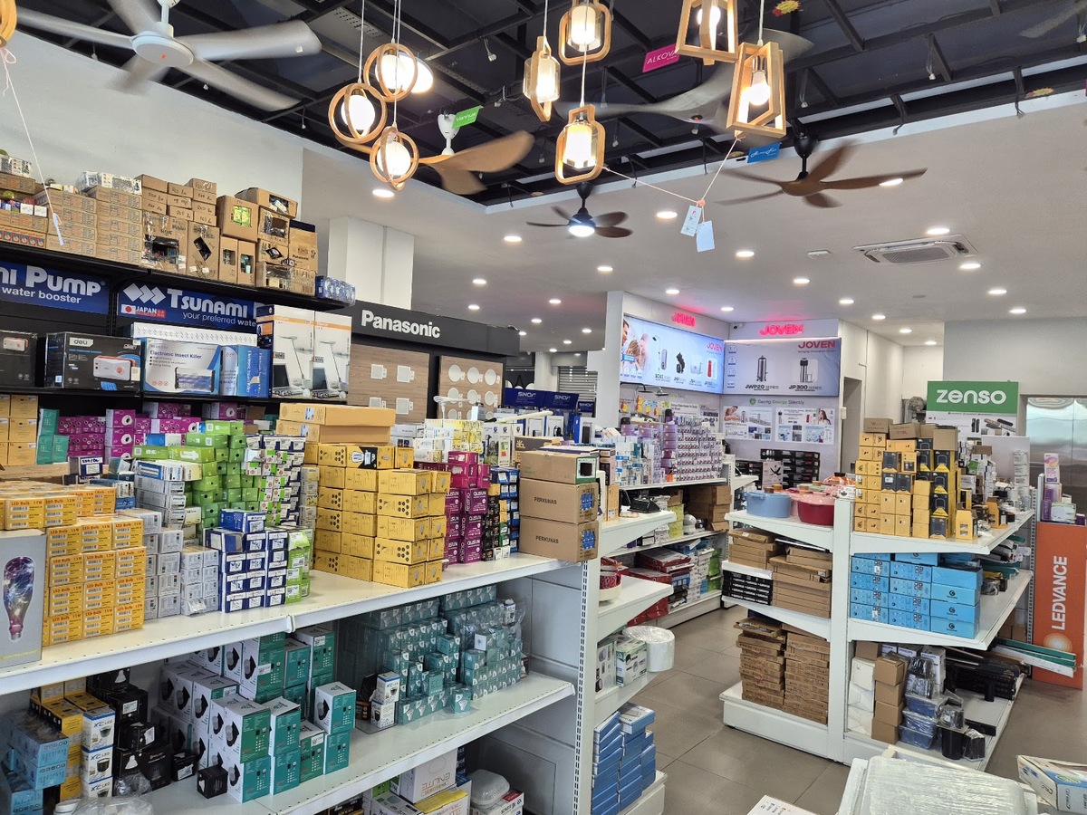 Store interior with white shelves stacked with colourful boxed bulbs and electrical accessories, ceiling fans and wooden pendant lamps overhead, and brand displays for Panasonic, Joven, Zenso and Ledvance