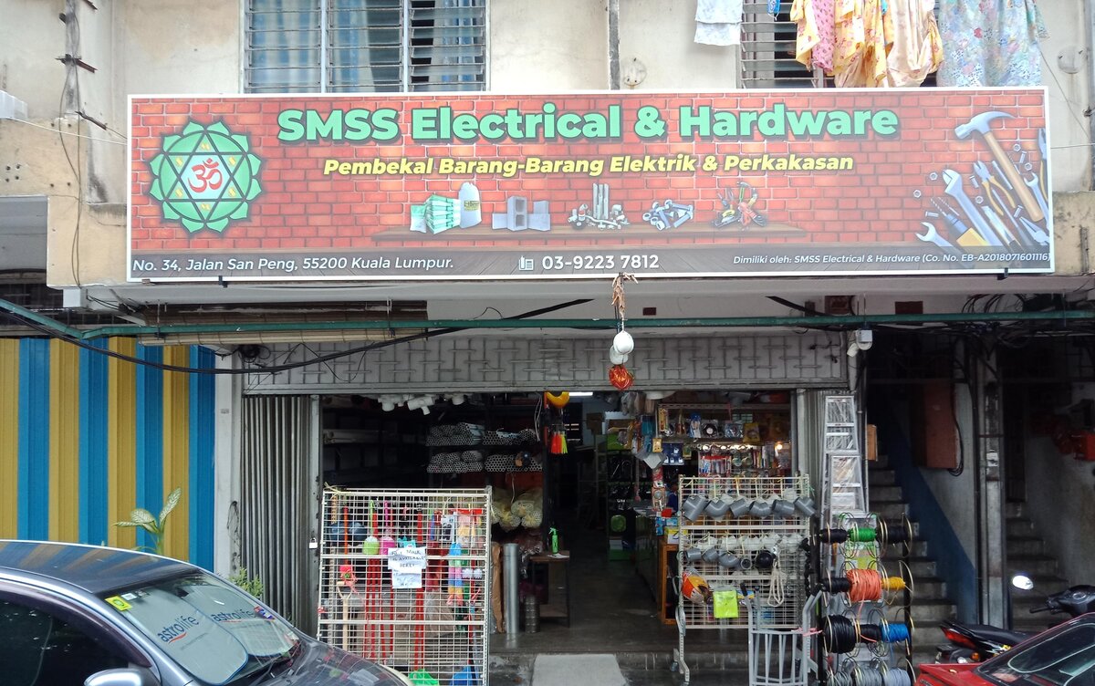 Red brick-pattern signboard reading SMSS Electrical & Hardware over an open shopfront with racks of pipe fittings, brooms and a stand of coloured reels