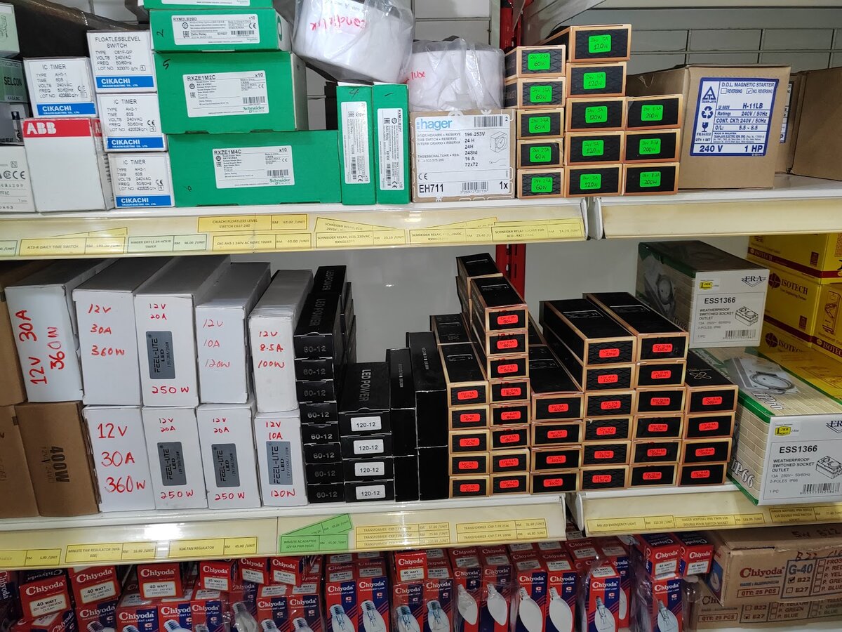 Shop shelves with green Schneider boxes, a Hager timer box, white Cikachi timer boxes, stacks of LED driver boxes and rows of Chiyoda lamp packs