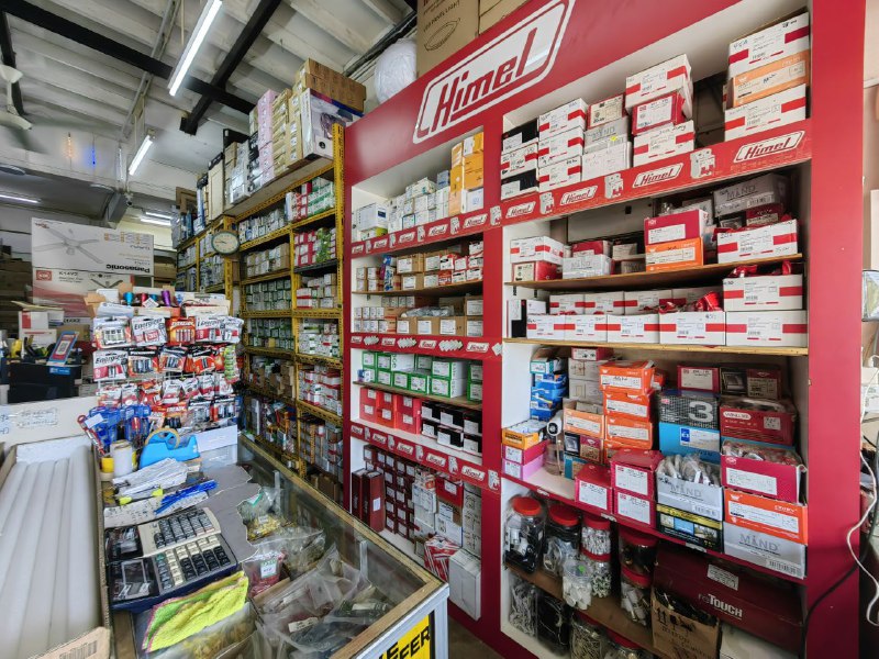 Narrow shop interior with a tall red shelving unit branded Himel full of small boxes, yellow shelves of boxed goods behind, and a glass counter in the foreground