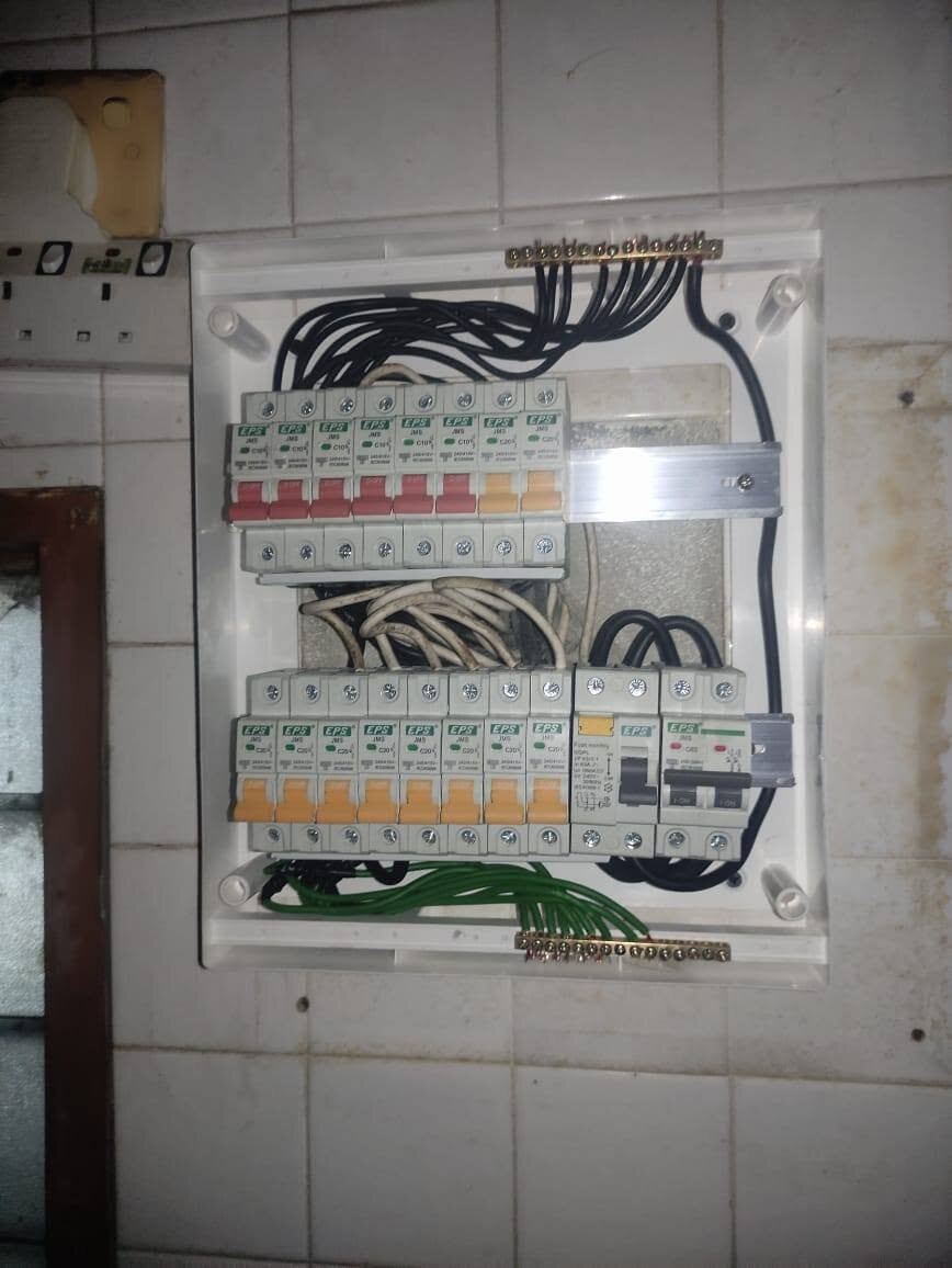 White DB box with a clear cover on a tiled wall: two rows of eight EPS breakers with red and orange toggles, an RCCB and a main switch on the right, black and green cables to brass bars at top and bottom, an old socket plate to the left