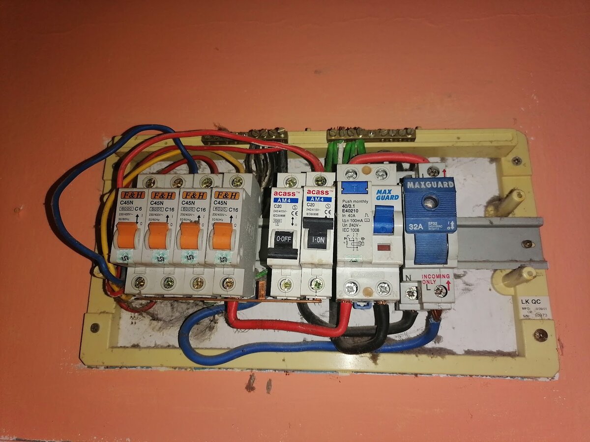 Opened surface-mounted distribution board on an orange wall, with four orange-handled MCBs, two black-handled MCBs, an RCCB with a blue test button and a blue main switch, wired in red, black and blue cable