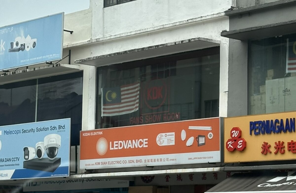 Row of shophouses; in the middle an orange Ledvance signboard over a shop, a KDK logo and Malaysian flag in the first-floor window, a blue CCTV company sign to the left and a yellow electrical shop sign to the right