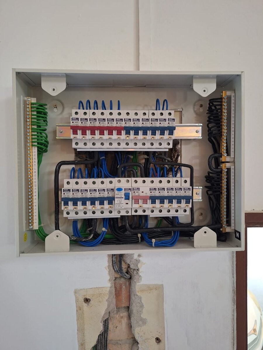 Recessed grey distribution board with two rows of red and blue Maxguard miniature circuit breakers, neat blue wiring, green earth wires and black neutral wires to bars on each side, above a freshly chased wall
