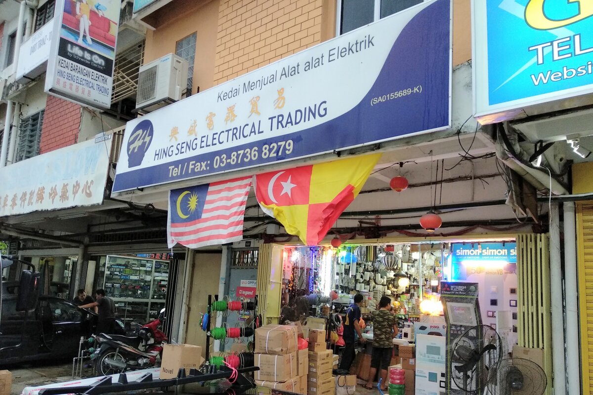 Shopfront with a white and blue signboard reading Hing Seng Electrical Trading and 03-8736 8279 above an open shop with cable reels, boxes, lamps and a Simon switch display, with Malaysian and Selangor flags hanging under the sign