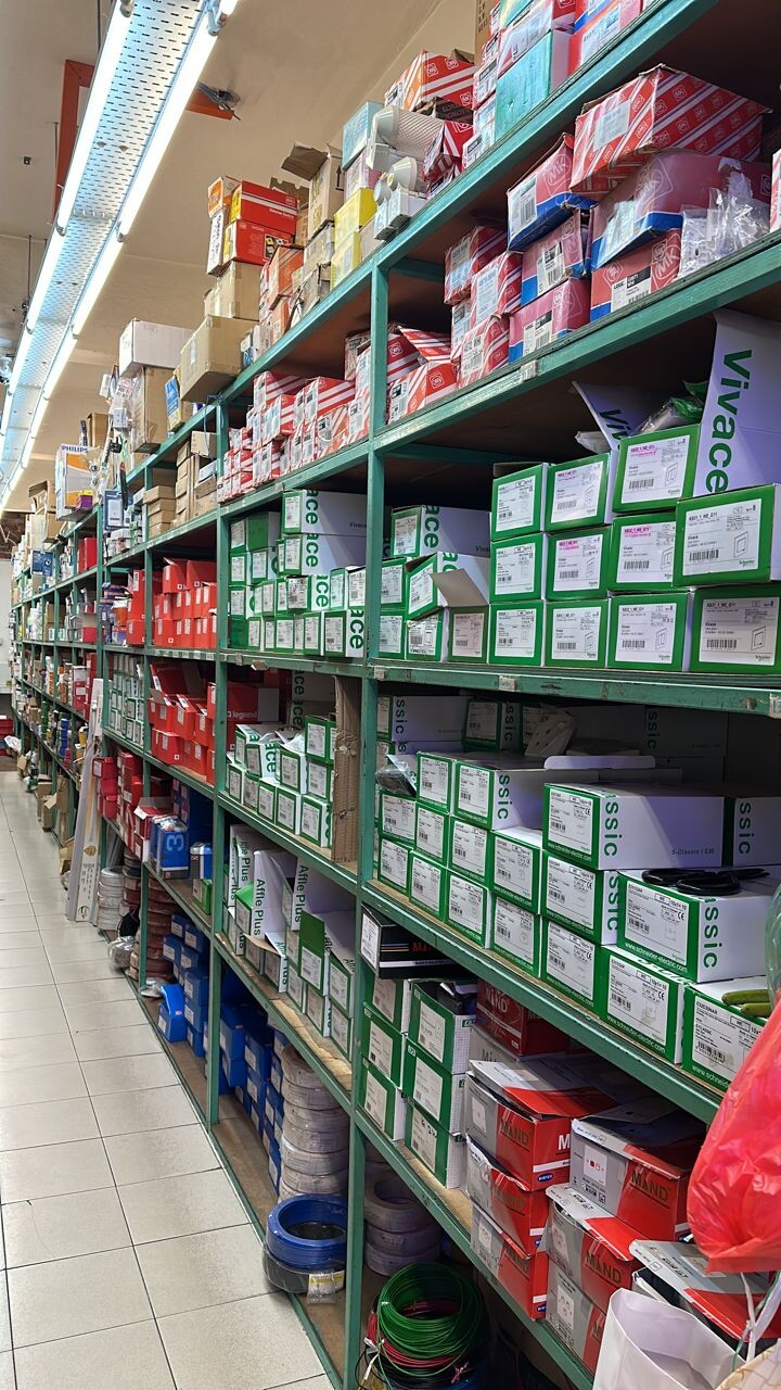 Aisle of green metal shelves stacked with red-and-white and green-and-white switch boxes, with coils of blue, grey and green cable on the bottom shelves