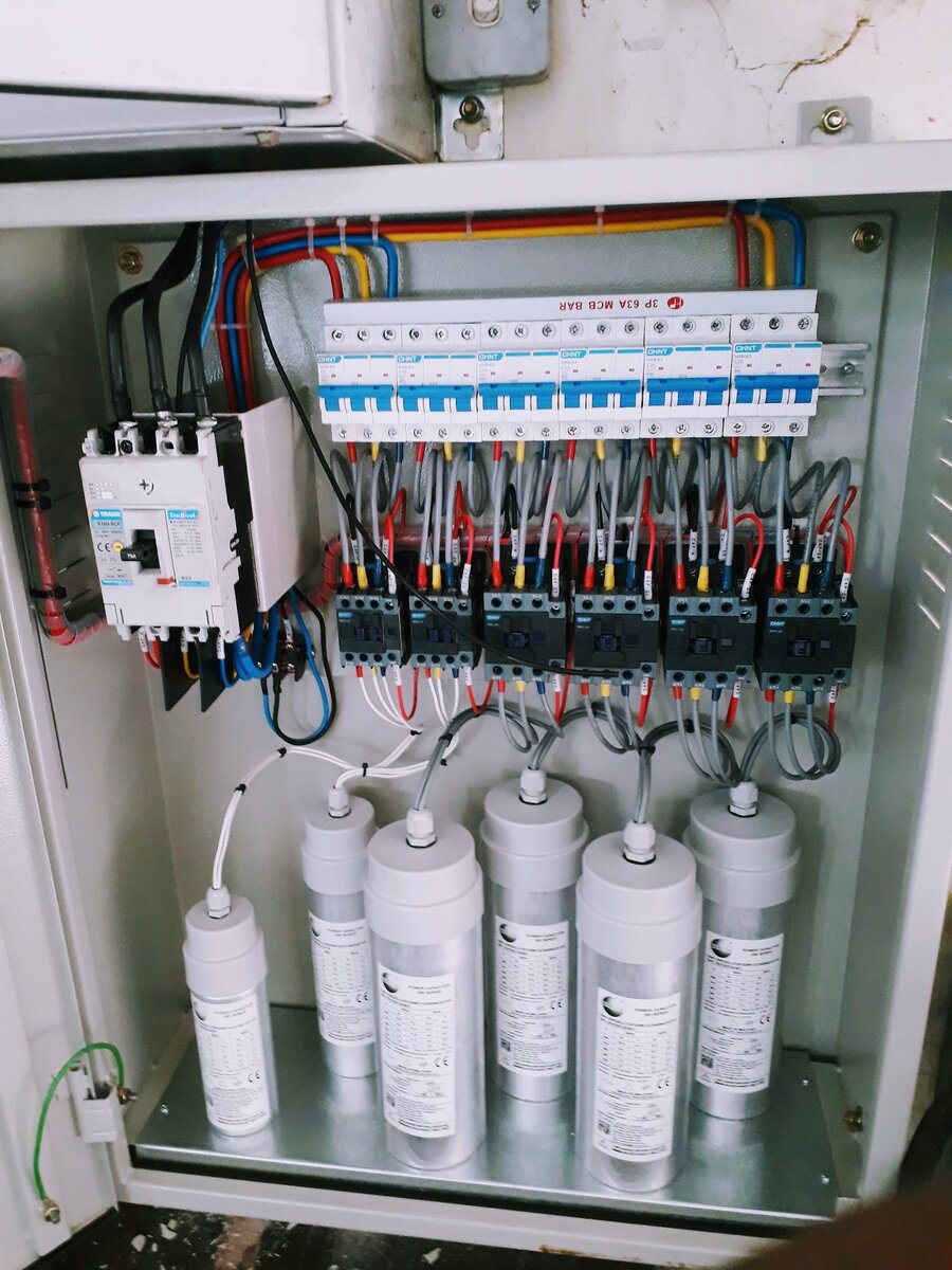 Open steel panel with a moulded-case circuit breaker, a row of blue-labelled MCBs, black contactors and six cylindrical capacitors at the bottom