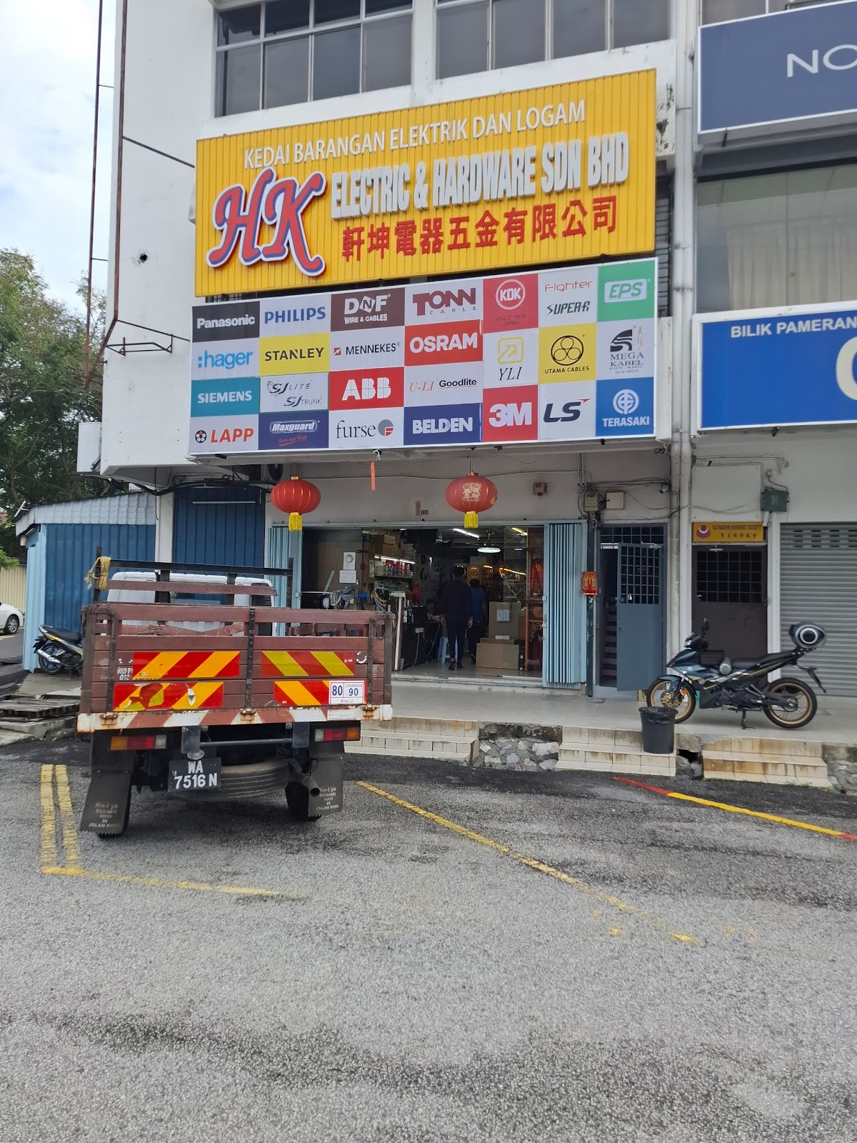 Yellow HK Electric & Hardware signboard above a large board of electrical brand logos at a corner shop