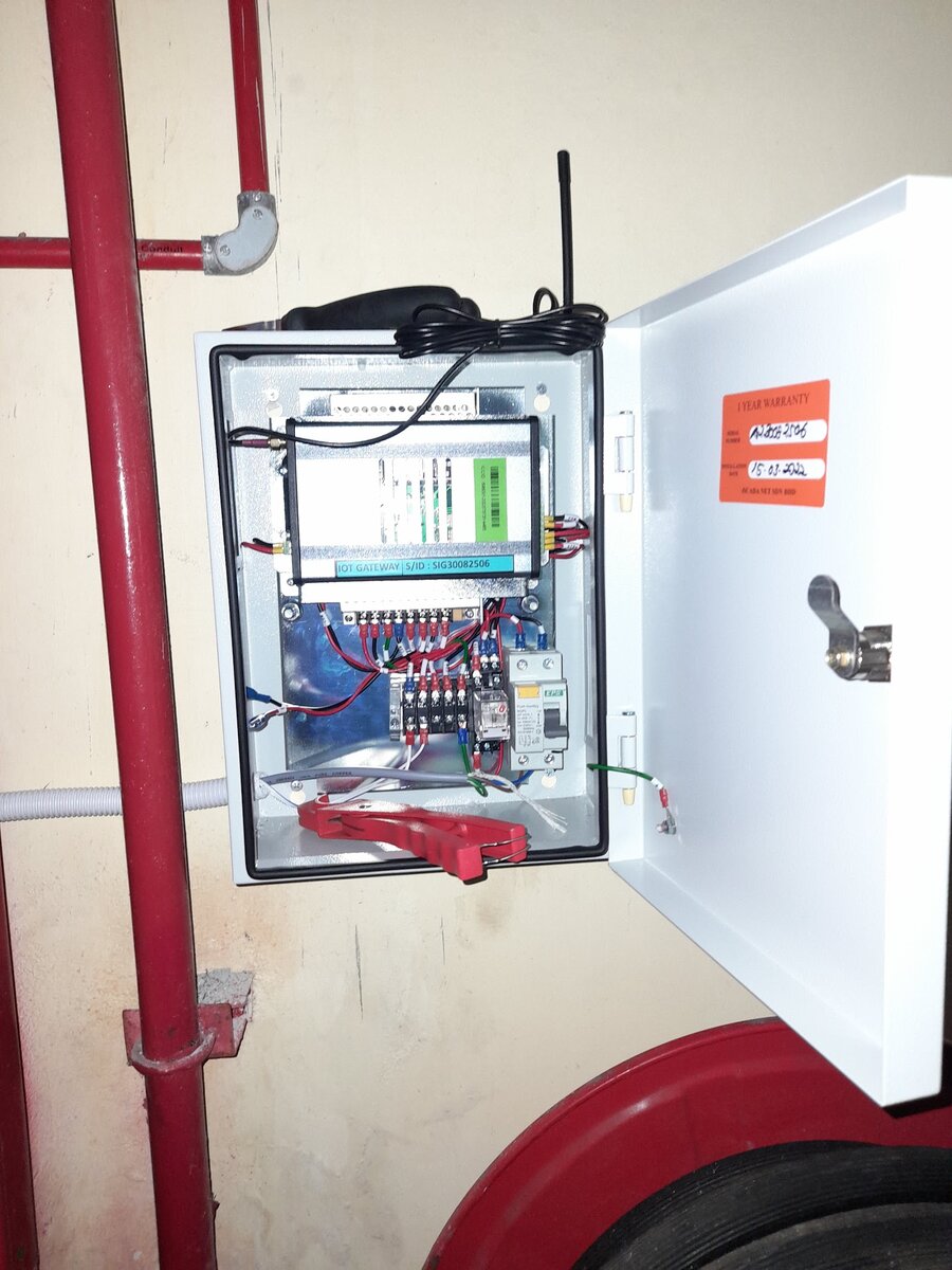 Open wall-mounted control panel with an antenna and breakers next to red pipes and a hose reel