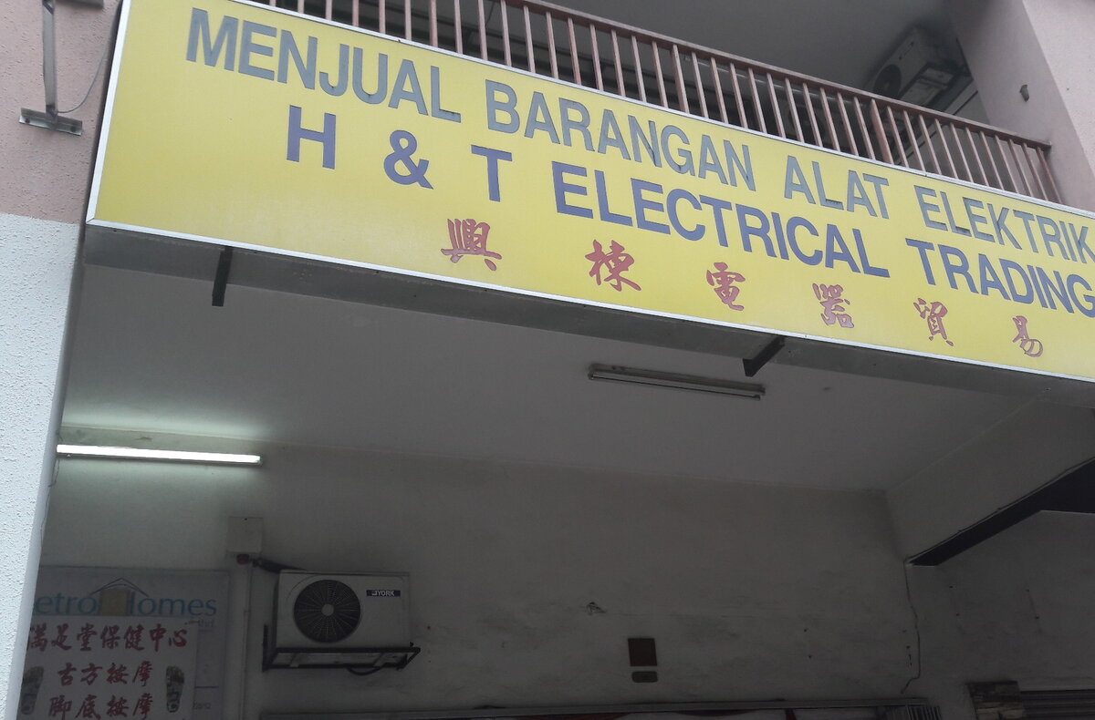 Yellow signboard reading Menjual Barangan Alat Elektrik, H & T Electrical Trading and Chinese characters, above a shuttered shop under a block of flats