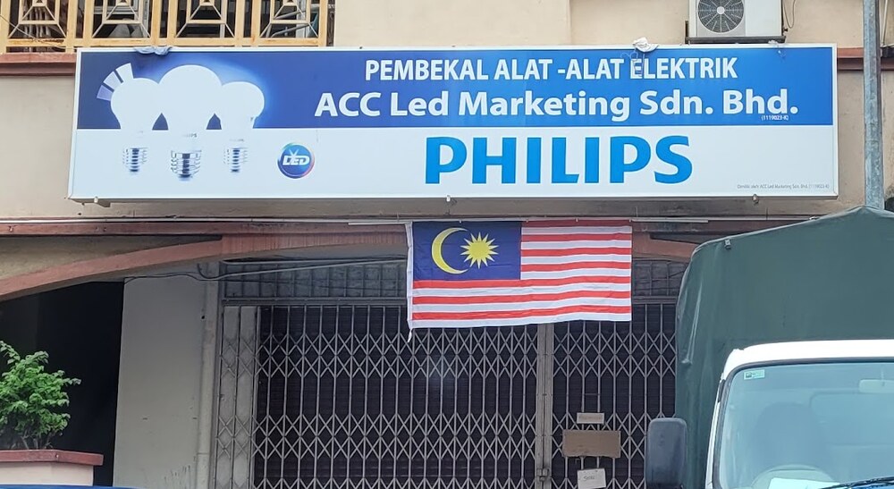 Blue and white signboard reading Pembekal Alat-Alat Elektrik ACC Led Marketing Sdn Bhd with a large Philips logo, above a closed grille shutter with a Malaysian flag
