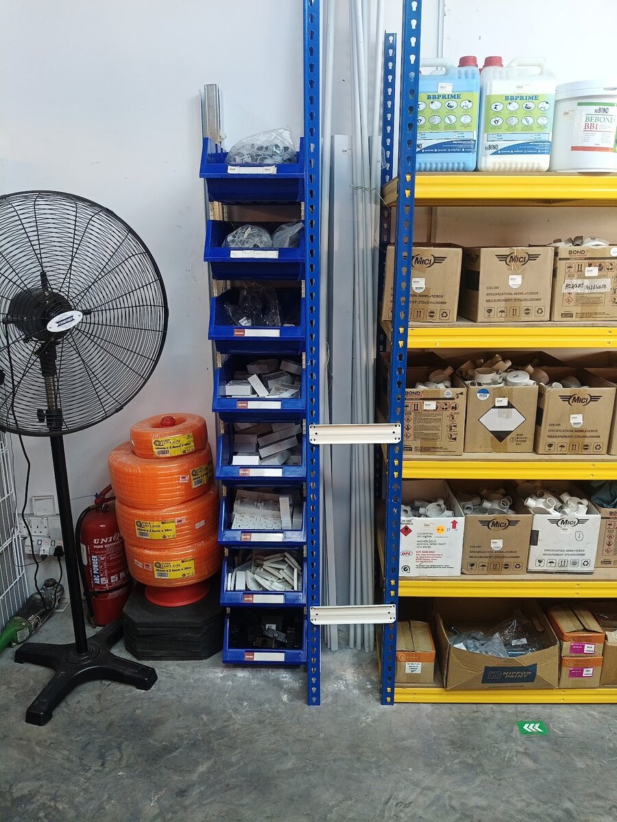 Blue stacking bins holding white PVC surface boxes and trunking fittings, with grey conduit lengths standing beside them, a stand fan and orange garden hose coils