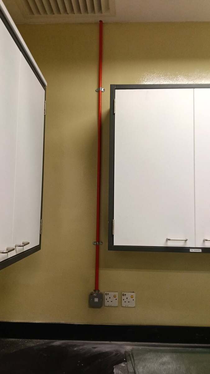 Yellow wall between two white wall cabinets with a vertical orange conduit, held by saddle clips, running from the ceiling down to a grey metal box and two white socket outlets near a dark worktop