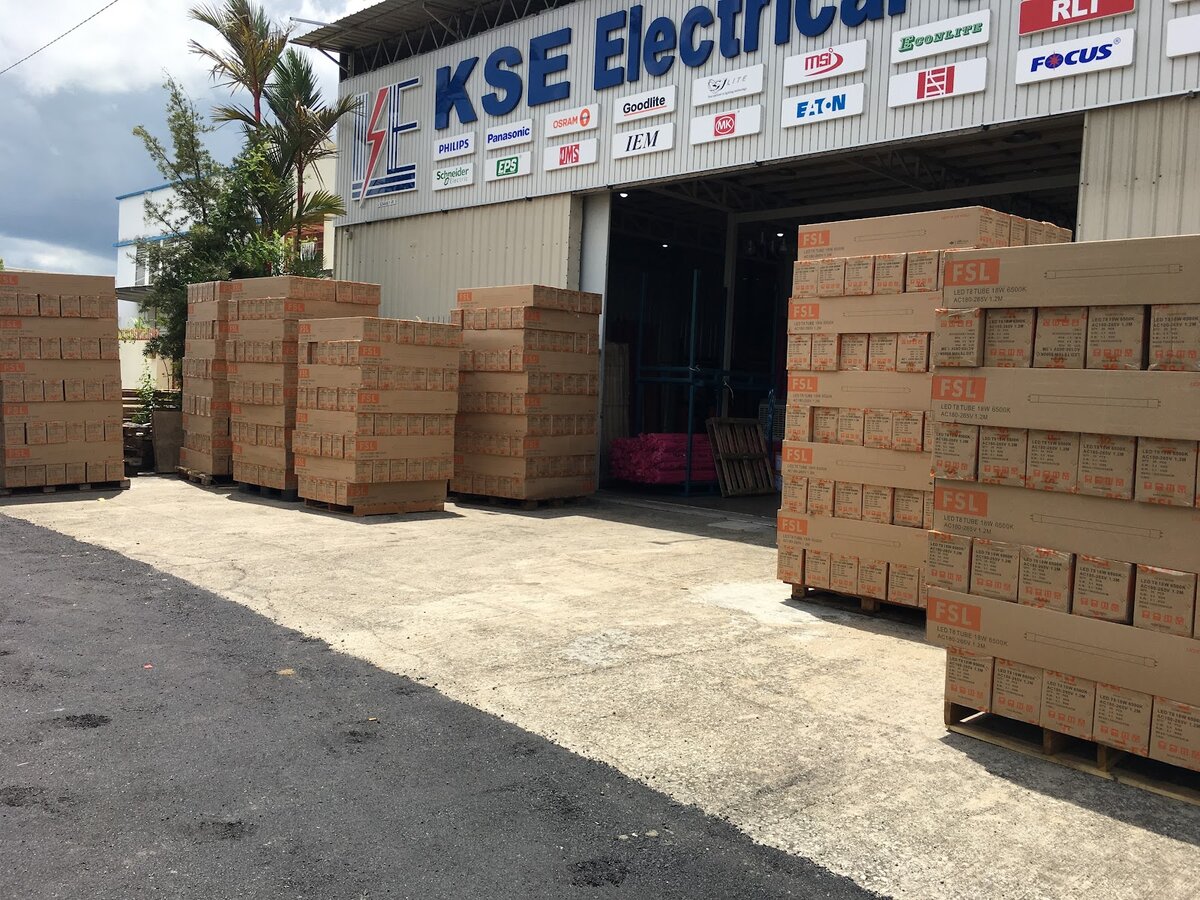 Warehouse front with large blue KSE Electrical lettering, rows of brand logo plates and stacked pallets of orange-labelled cartons