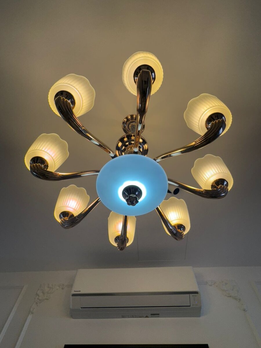 Brass chandelier with eight glass shades on a white ceiling, an air-conditioner unit on the wall below