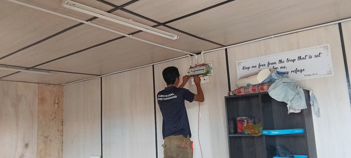 Worker in a navy shirt fitting a small breaker board high on a laminate wall, with white conduit running along the ceiling to fluorescent lights