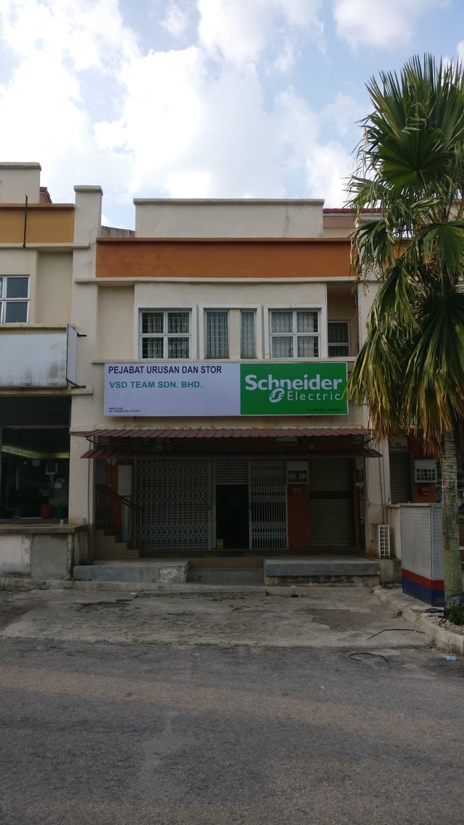 Two-storey shop lot with a white and green signboard reading Pejabat Urusan dan Stor, VSD Team Sdn. Bhd. and Schneider Electric, above a closed grille