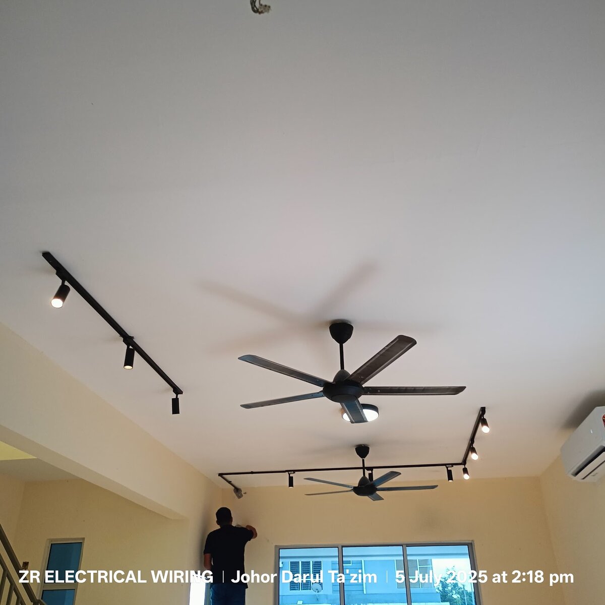Living room with two black ceiling track-light rails, two ceiling fans and a technician working on a wall near the window