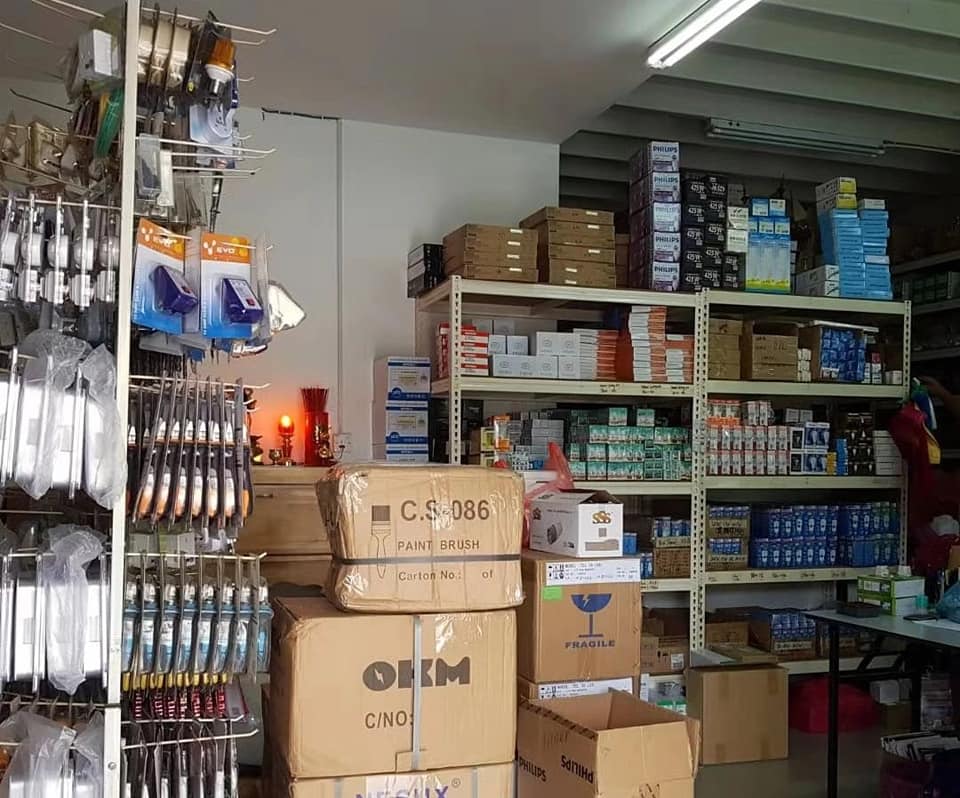 Room with metal shelves stacked with boxed lamps, cartons on the floor and a hanging rack of tools on the left