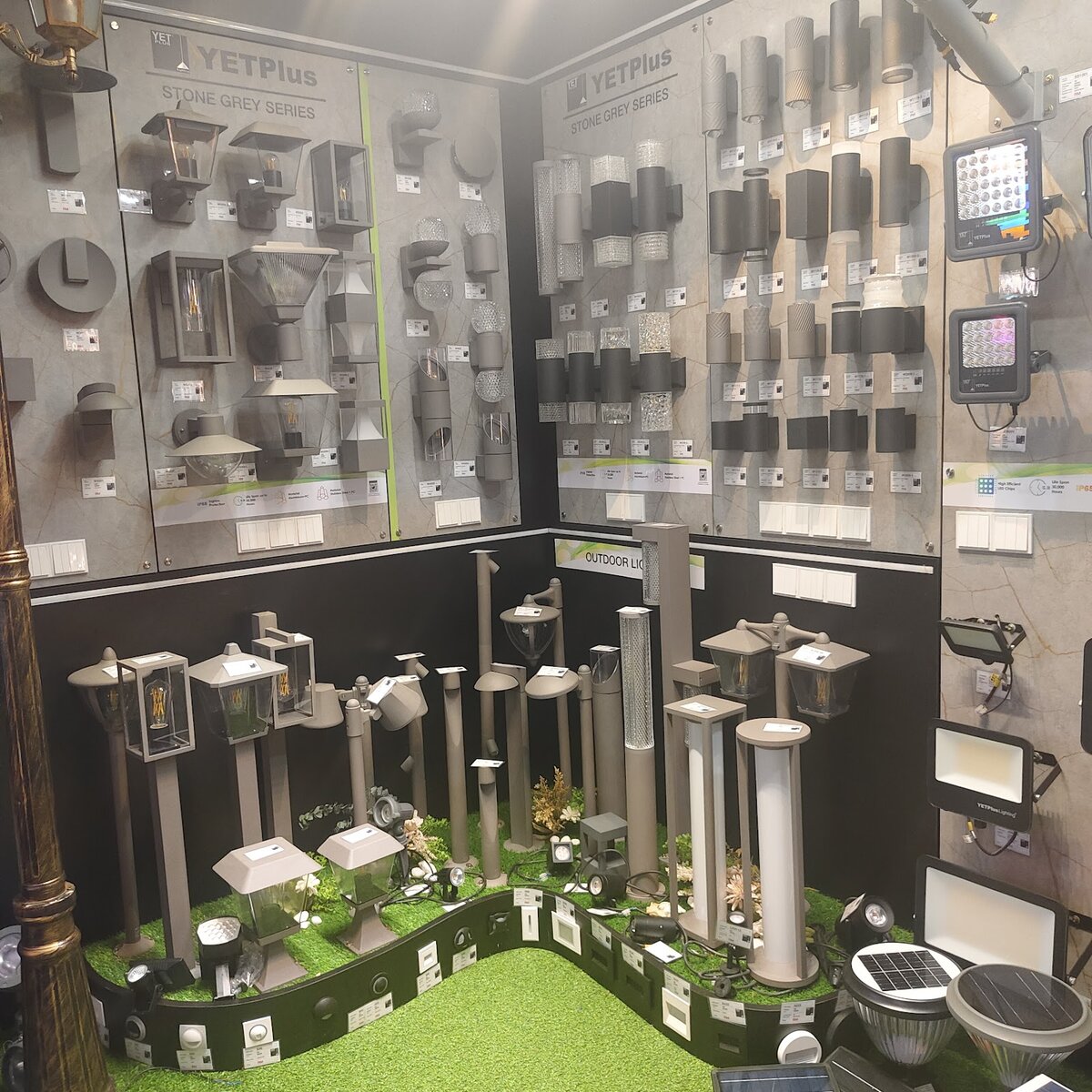 Corner display of grey and black outdoor wall lamps above a bed of artificial grass with garden bollard lights