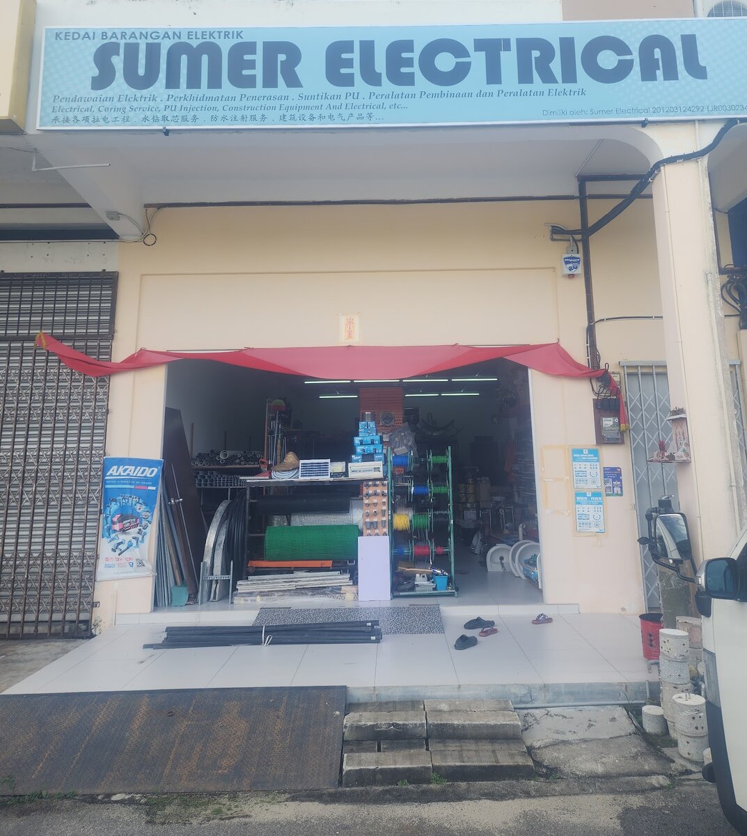 Shophouse with a light-blue "Kedai Barangan Elektrik Sumer Electrical" signboard above an open shopfront with cable reels, pipes, green mesh and a power-tool banner