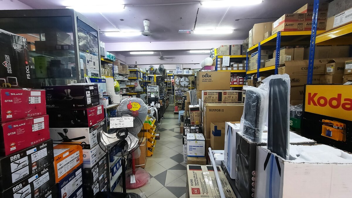 Shop aisle lined with stacked ceiling-fan cartons, stand fans and shelves of boxed goods under fluorescent lights