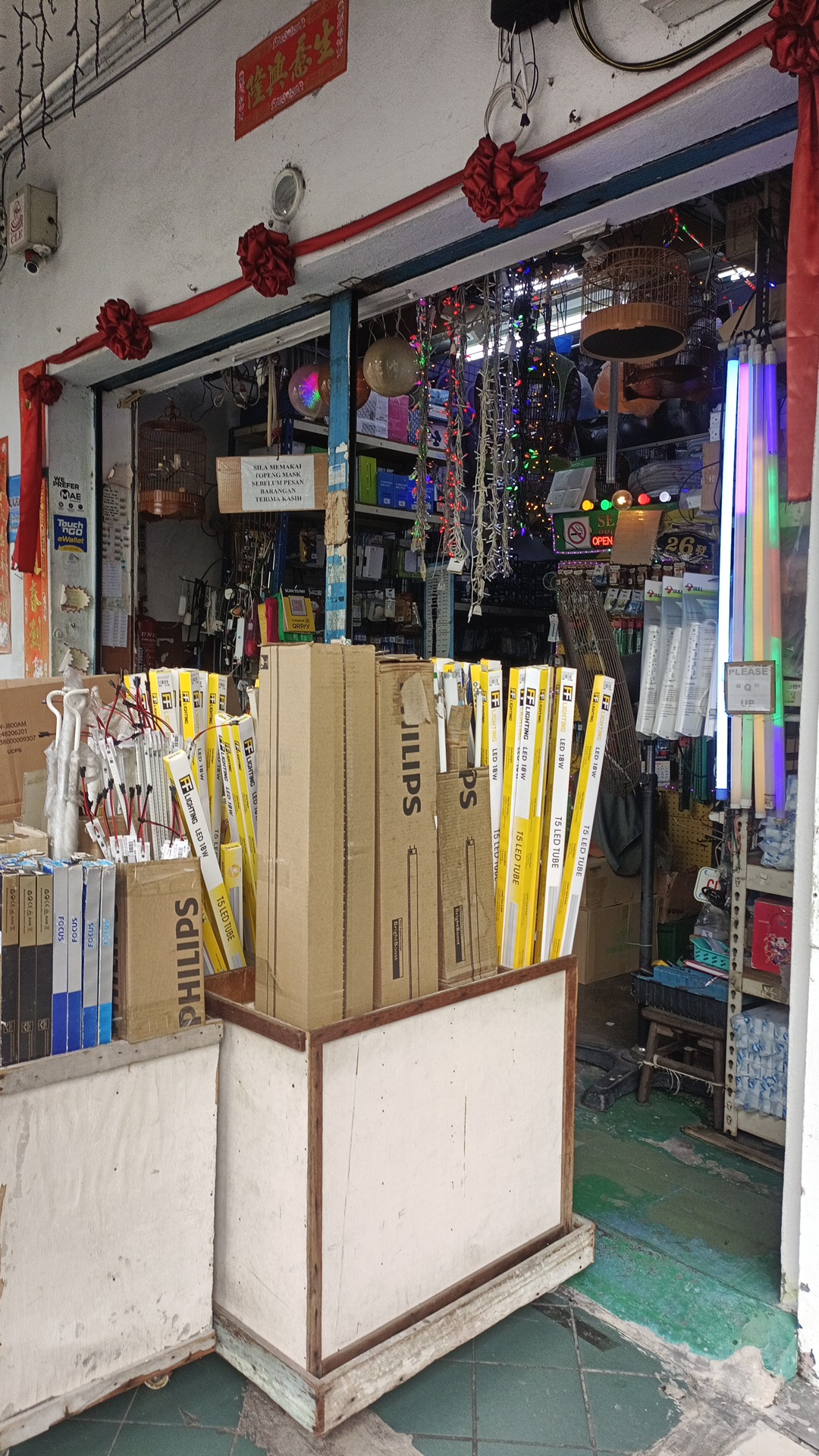 Open shopfront of an electrical shop with cartons marked Philips and yellow FF Lighting T5 LED tube packs in bins at the entrance, and shelves of electrical goods inside