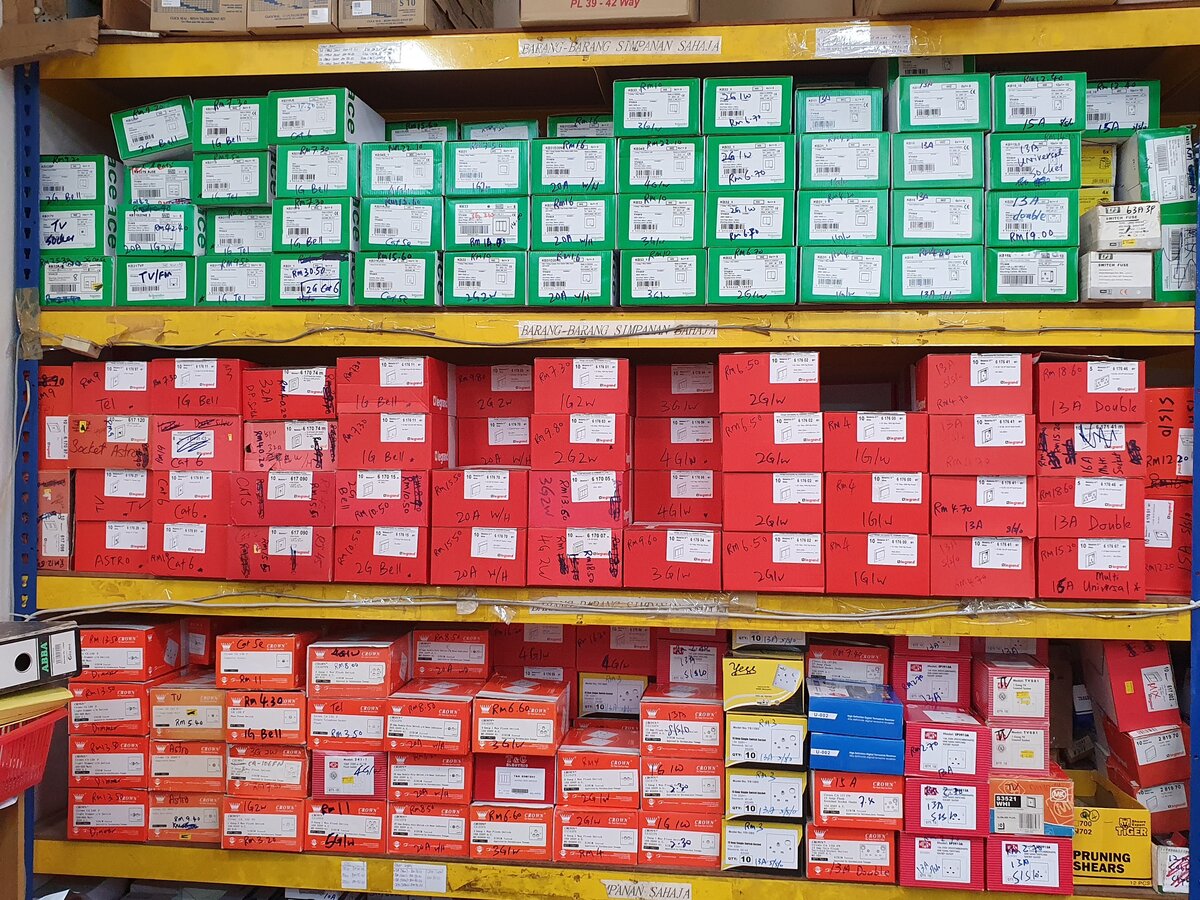 Yellow storage racks packed with green Schneider and red Legrand and Crown switch and socket boxes with handwritten labels and prices