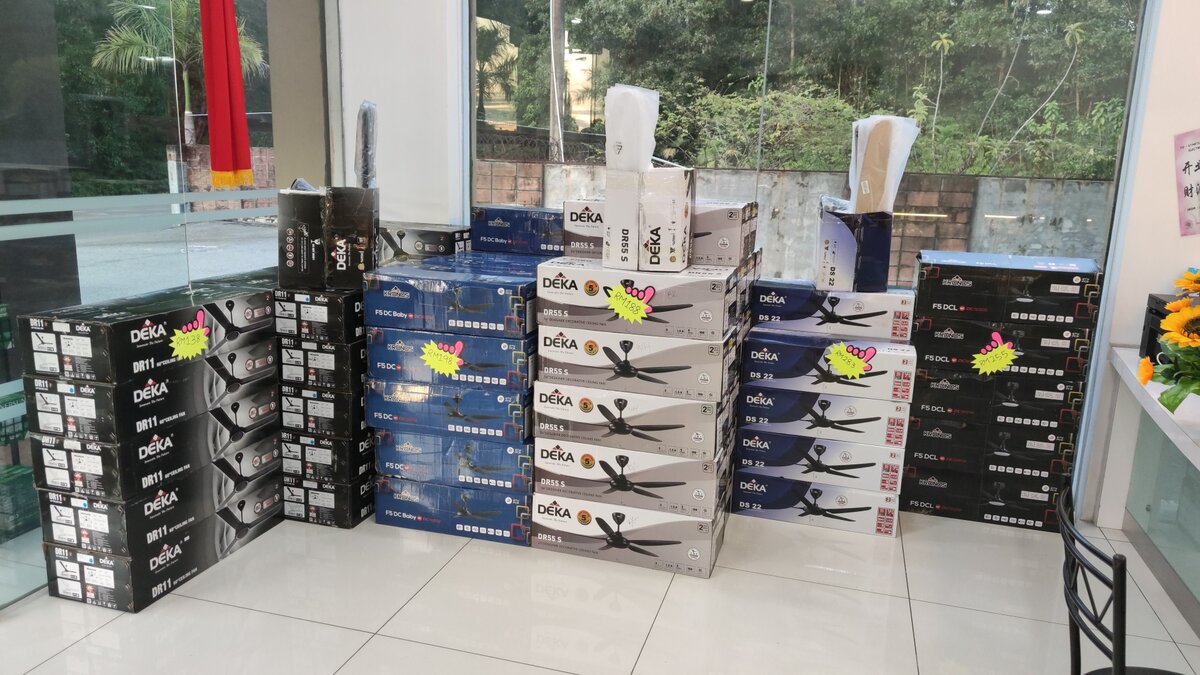 Stacks of black, blue and white ceiling-fan boxes labelled Deka and Kronos with pink price stickers by a glass shop window