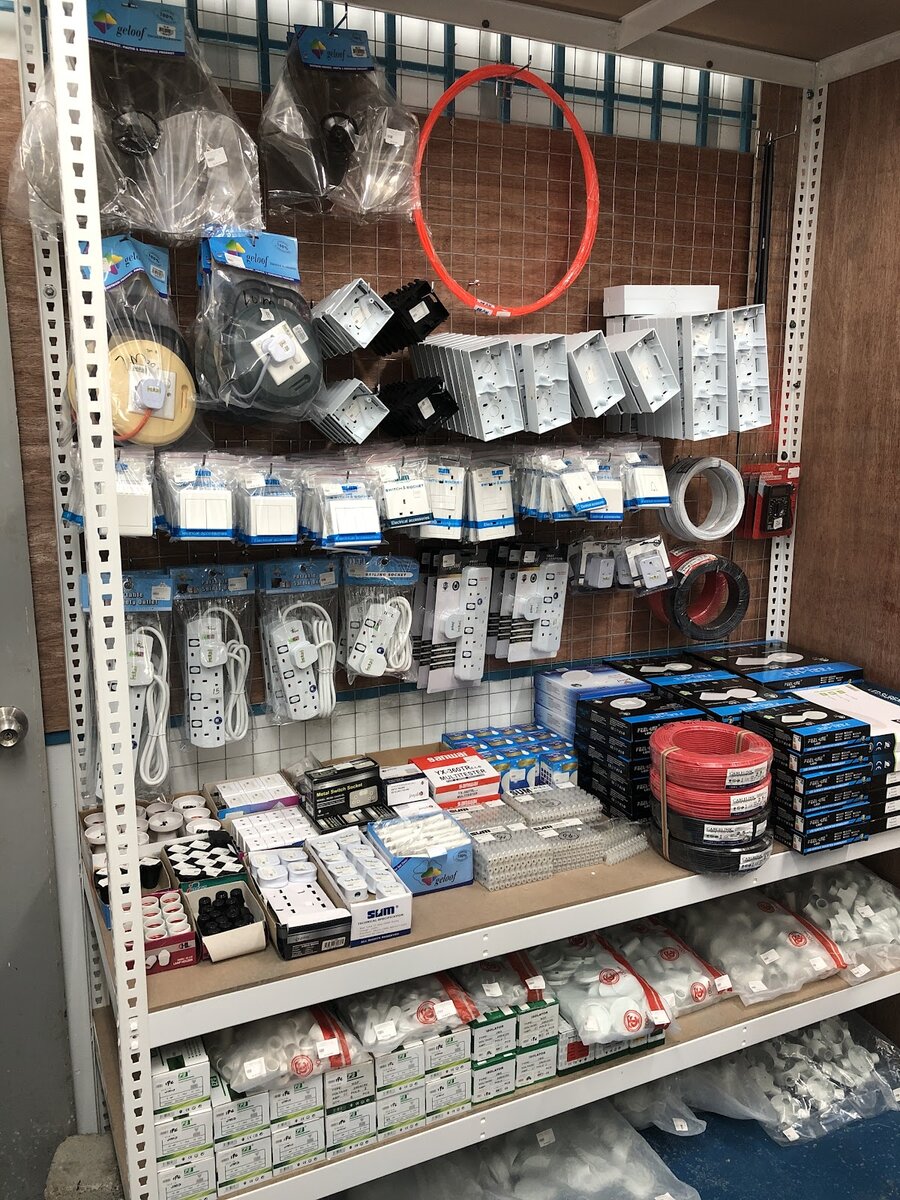 Shelving bay with packed switch sockets, surface boxes, cable reels and extension sockets on a wire grid, and boxed LED downlights, red and black wire coils and conduit fittings on the shelves