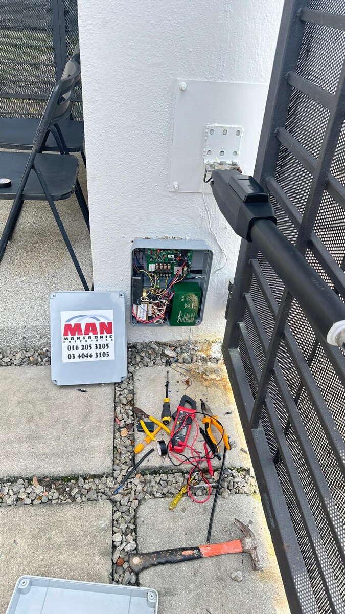 An opened grey autogate controller box on a house wall with a green circuit board and transformer, its cover on the ground below carrying a red and white MAN Mantronic Enterprise sticker with two phone numbers, tools on the paving