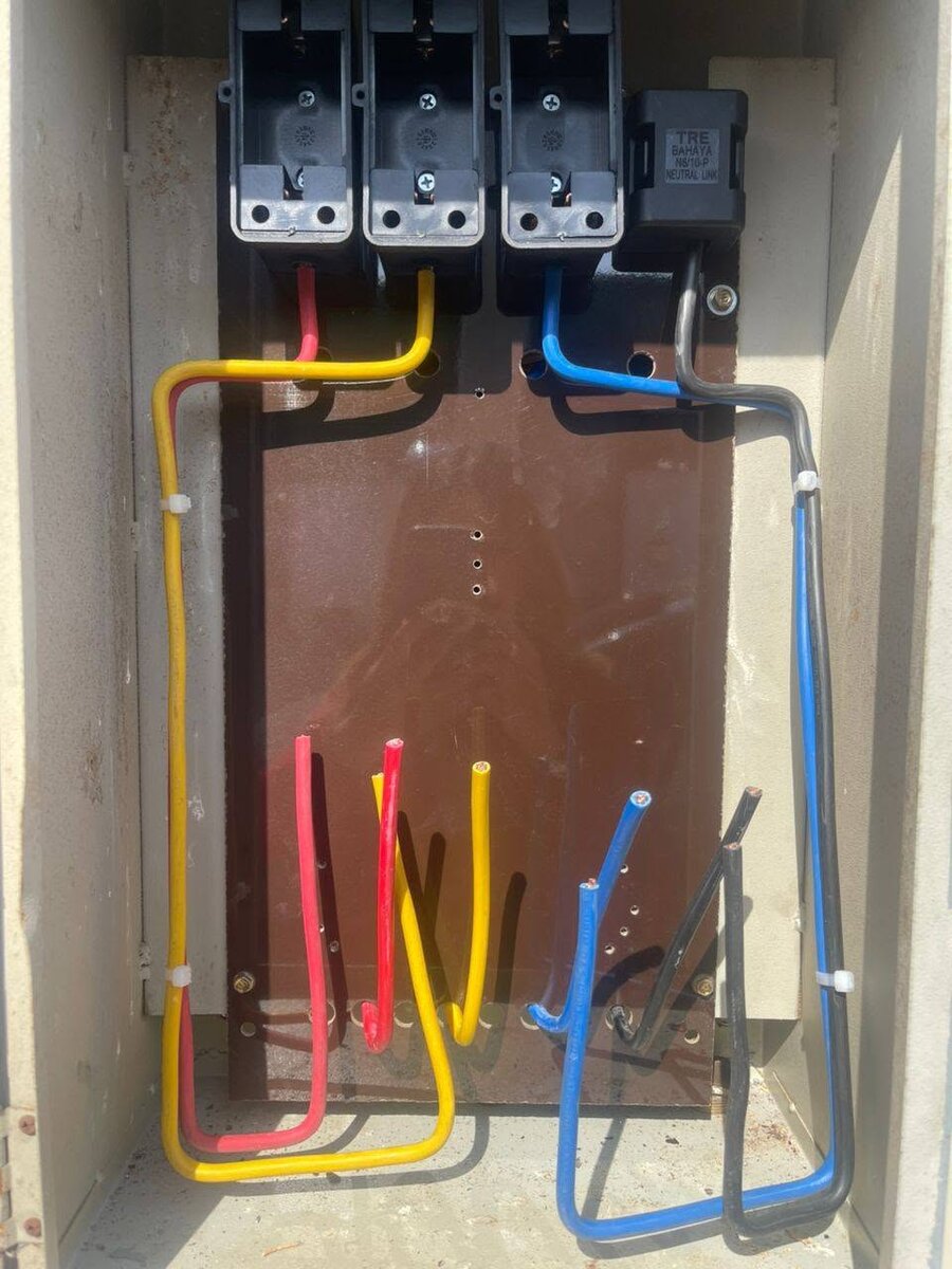 Open grey metal meter enclosure with three black fuse-carrier bases and a black neutral link at the top of a brown board, red, yellow, blue and black cables looped below with loose ends