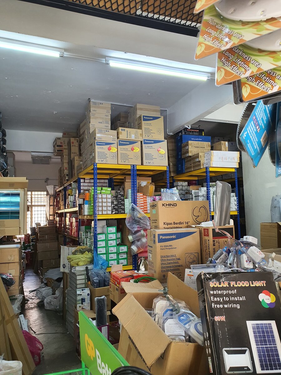 Crowded shop with blue and yellow racks stacked with boxed Panasonic, KDK and Khind fans, small boxed electrical parts and a solar flood light box