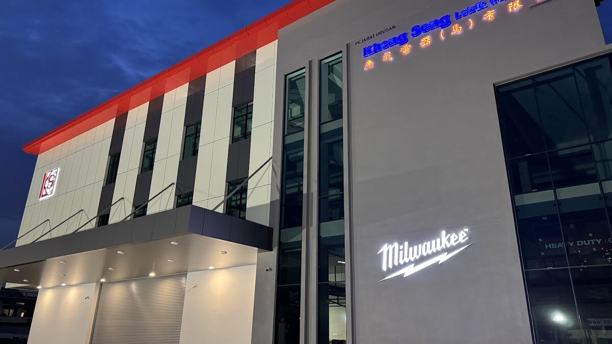 Modern three-storey building with grey and white panels, a red roof edge, a KS logo, a lit Milwaukee sign tall glass panels and a roller shutter under a canopy