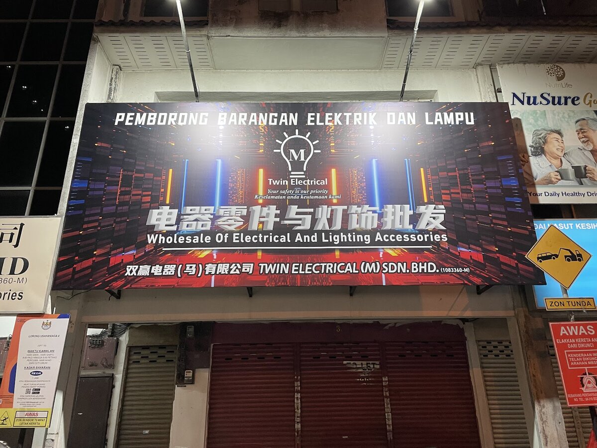 Dark signboard reading Pemborong Barangan Elektrik dan Lampu, Twin Electrical, Wholesale of Electrical and Lighting Accessories, above closed red shutters