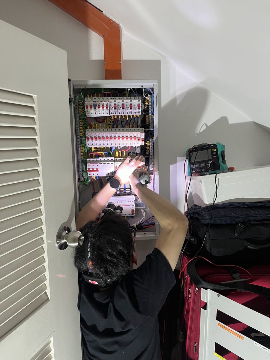 A technician with a head torch and a torch in hand reaching into an open distribution board fitted under a sloping staircase ceiling, with a tester on a white cabinet beside him
