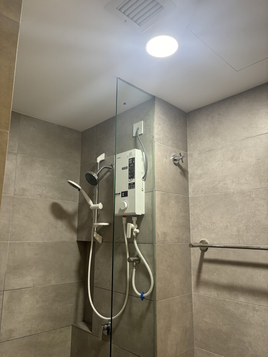 Instant water heater with a hand shower on a grey-tiled shower wall behind a glass screen, with a round ceiling light above
