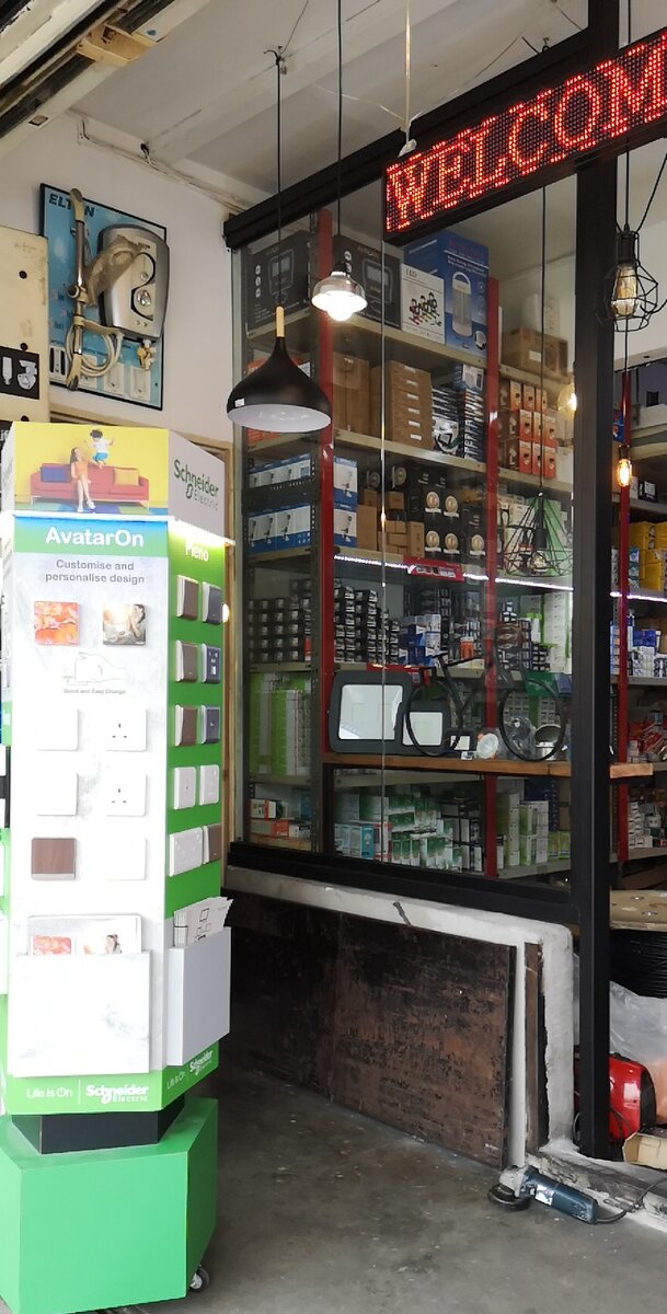 Shop entrance with a green-and-white Schneider AvatarOn switch display tower in front of shelves stacked with lamp boxes and hanging pendant lights
