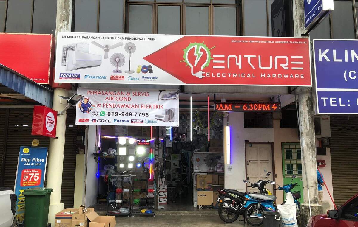 Shopfront with a red Venture Electrical Hardware signboard showing aircond and fan pictures, a white banner advertising aircond installation and electrical wiring, a rack of coloured cable reels, lit LED panel lights and fans inside