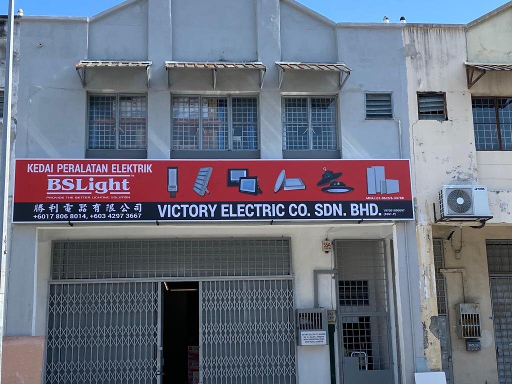 Two-storey grey shophouse with a red signboard reading Kedai Peralatan Elektrik, BSLight and Victory Electric Co. Sdn. Bhd. with pictures of LED street lights, floodlights, panel lights and high-bay lamps, above a half-open folding grille