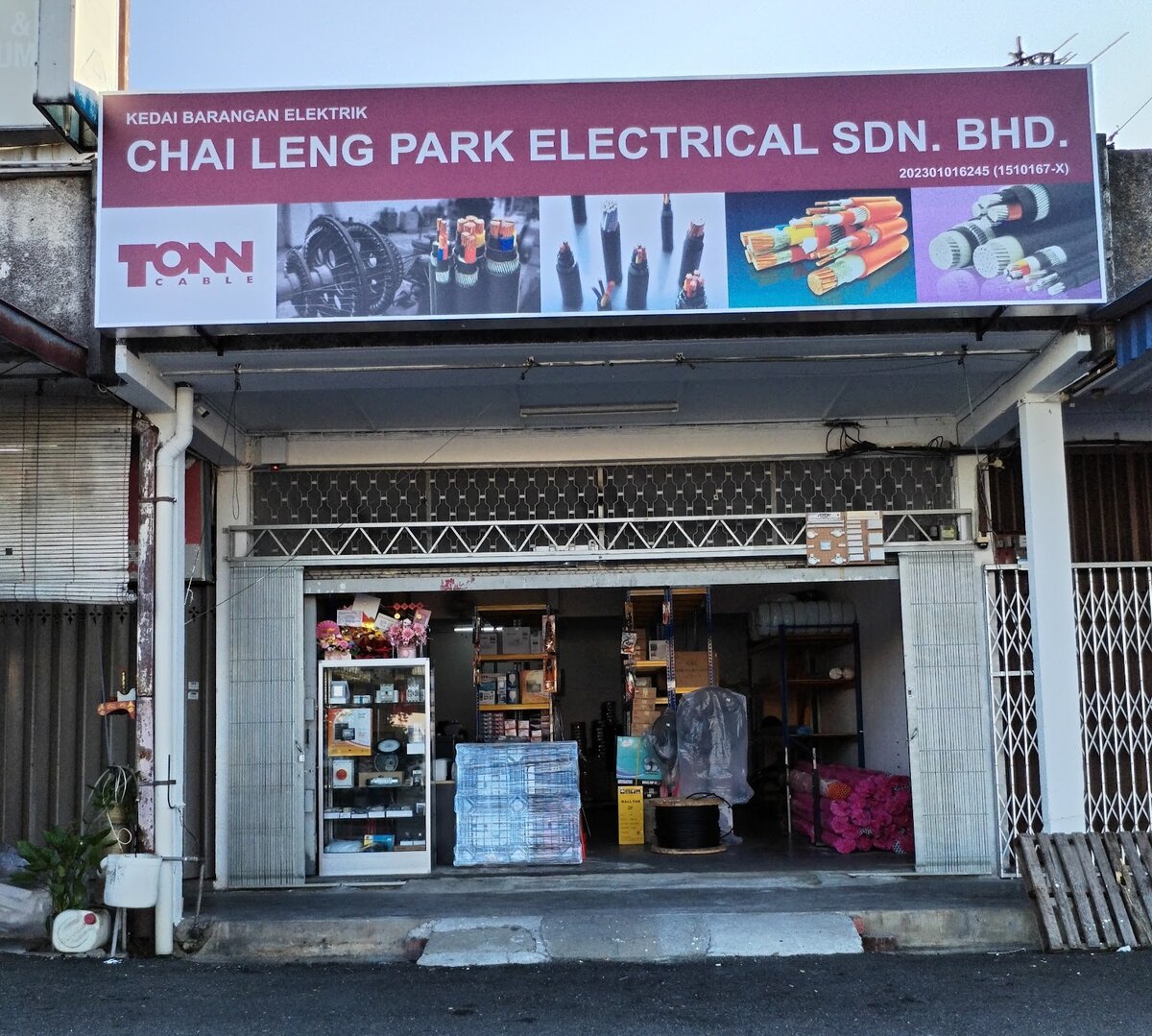 Maroon signboard reading Kedai Barangan Elektrik Chai Leng Park Electrical Sdn Bhd with a Tonn Cable logo and cable pictures, over an open shopfront with a glass display cabinet, a black cable drum and stacked cartons