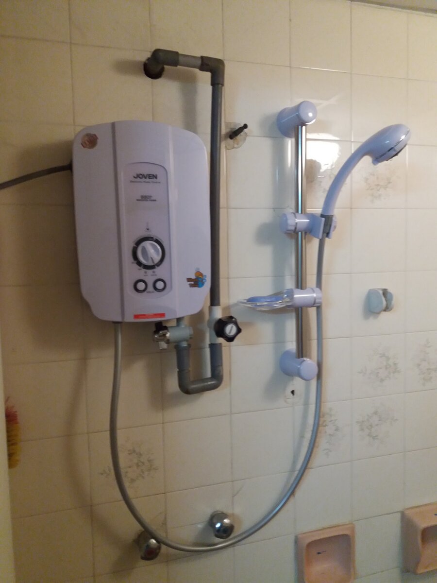 White Joven water heater with dial and buttons mounted on a tiled bathroom wall beside a shower rail and hand shower