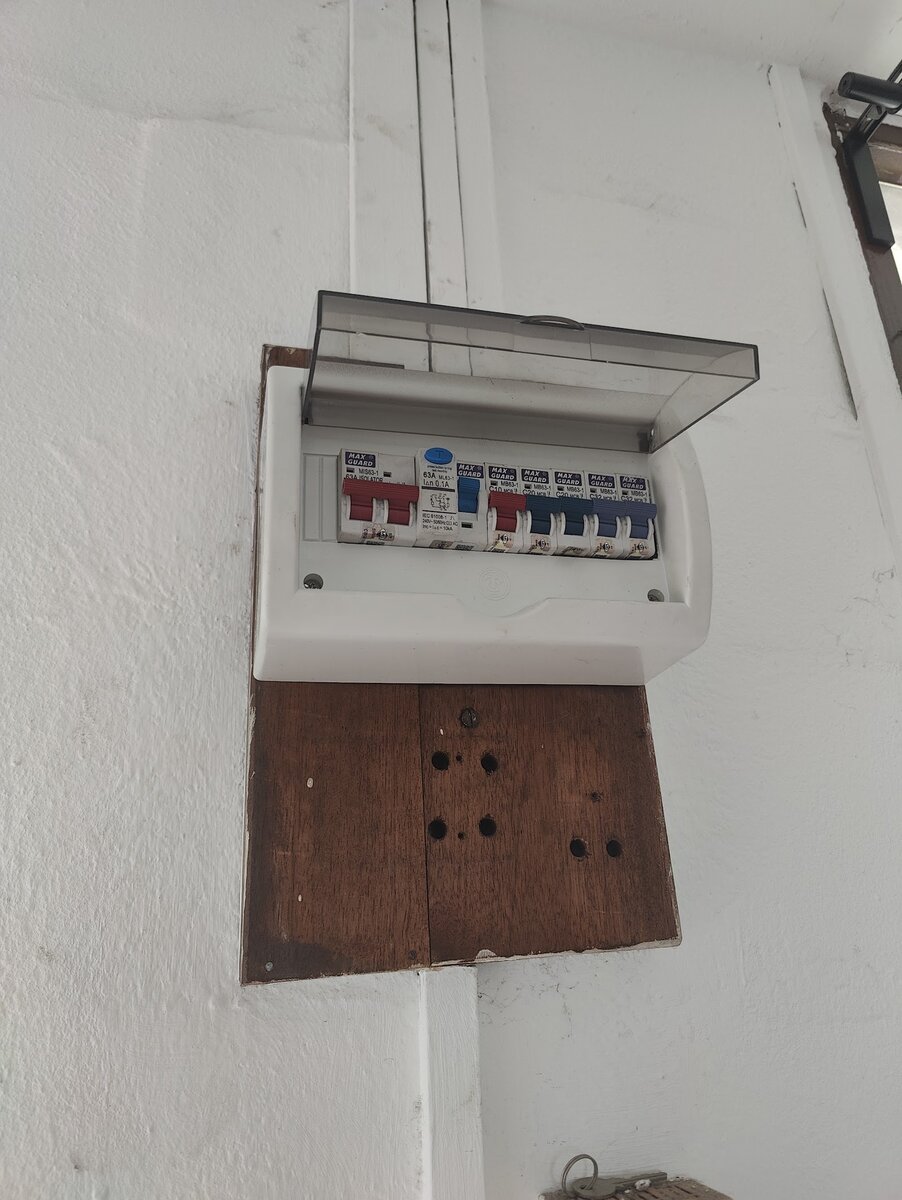 Small white distribution board with an open clear cover showing a red isolator, an RCCB and a row of miniature circuit breakers, mounted above an old brown wooden board with empty holes