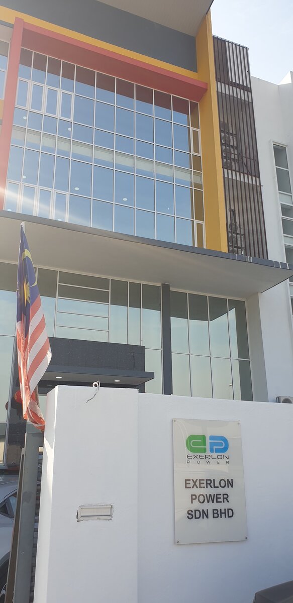 Three-storey glass-fronted factory office with red and yellow trim and an Exerlon Power Sdn Bhd plate on the gate pillar