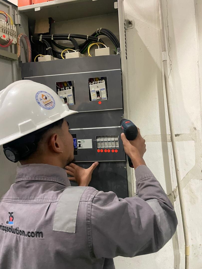 Technician in a white hard hat and grey uniform using a drill on an open electrical switchboard