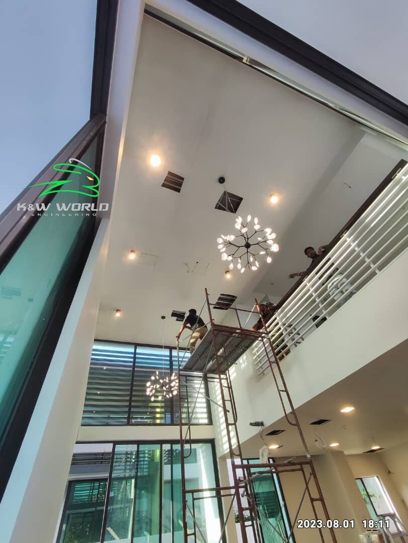 View up into a tall living space with recessed downlights and a branch chandelier, one worker on scaffolding and another at a gallery railing