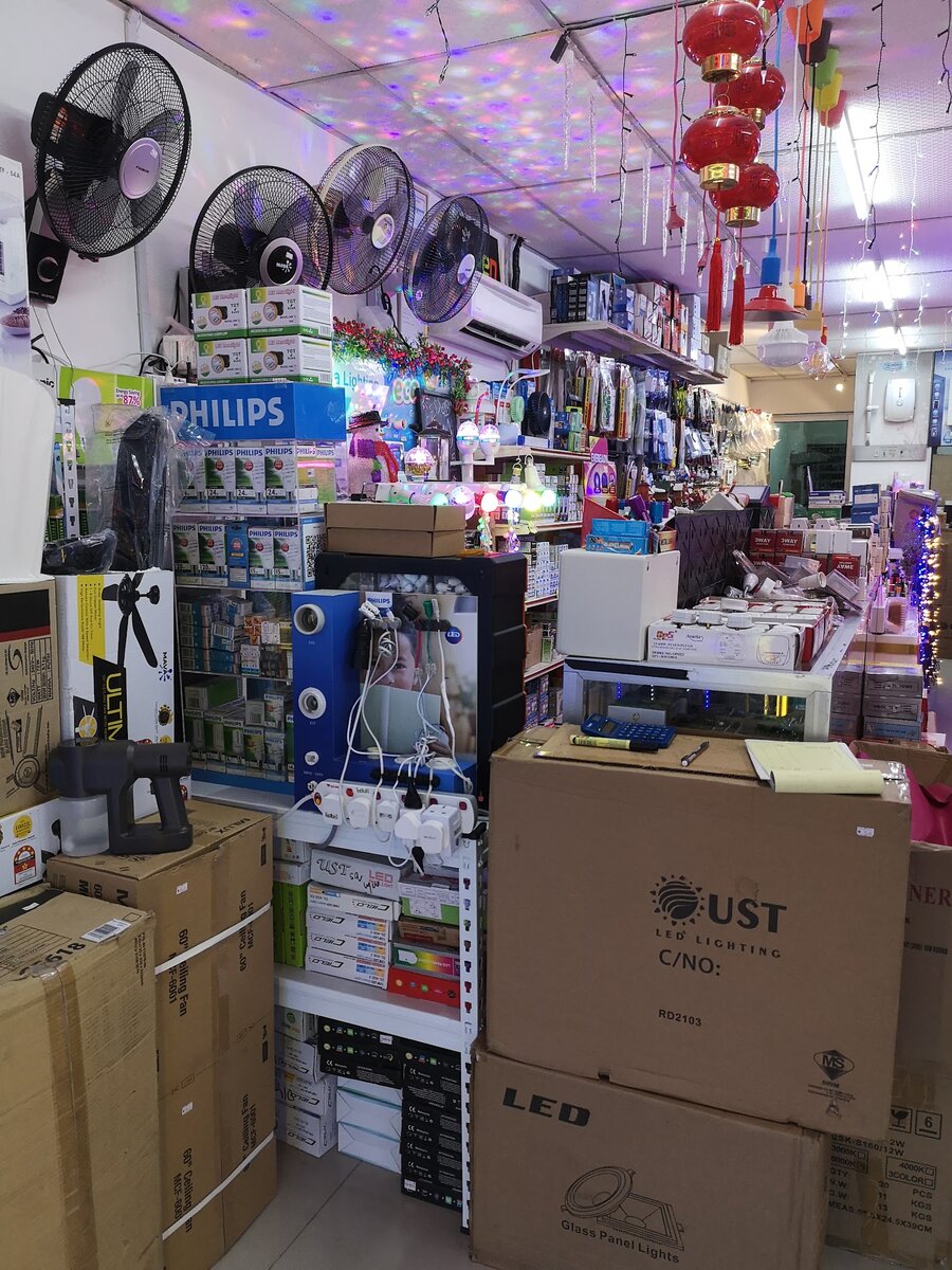 Crowded shop interior with wall fans on the left wall, a blue Philips lamp display stand, boxed LED panel lights in front, a counter with 3-way adaptors and fused plugs, and hanging pendant lamps