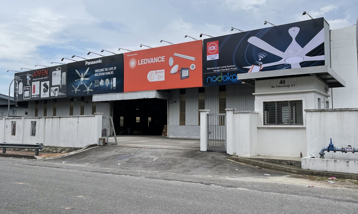 Low warehouse behind a white wall with four large billboards on the roof edge (Joven, Panasonic, orange Ledvance, KDK ceiling fan) and a gatehouse marked 40 Jln Teknologi 3/1